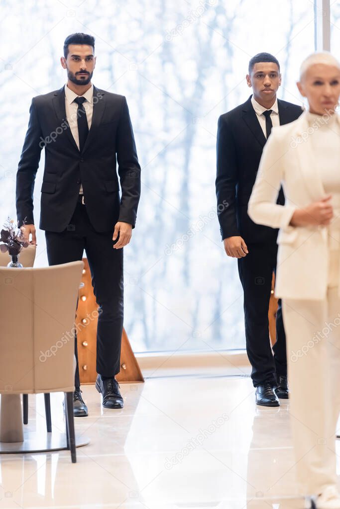 multiethnic bodyguards in suits walking in hotel foyer near senior businesswoman on blurred foreground