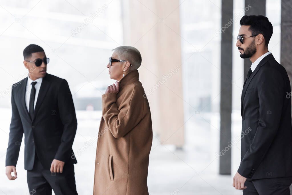 interracial guards in black suits protecting stylish senior businesswoman on street