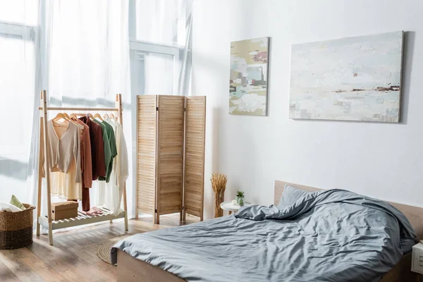 modern bed near folding screen and rack with clothes in bedroom