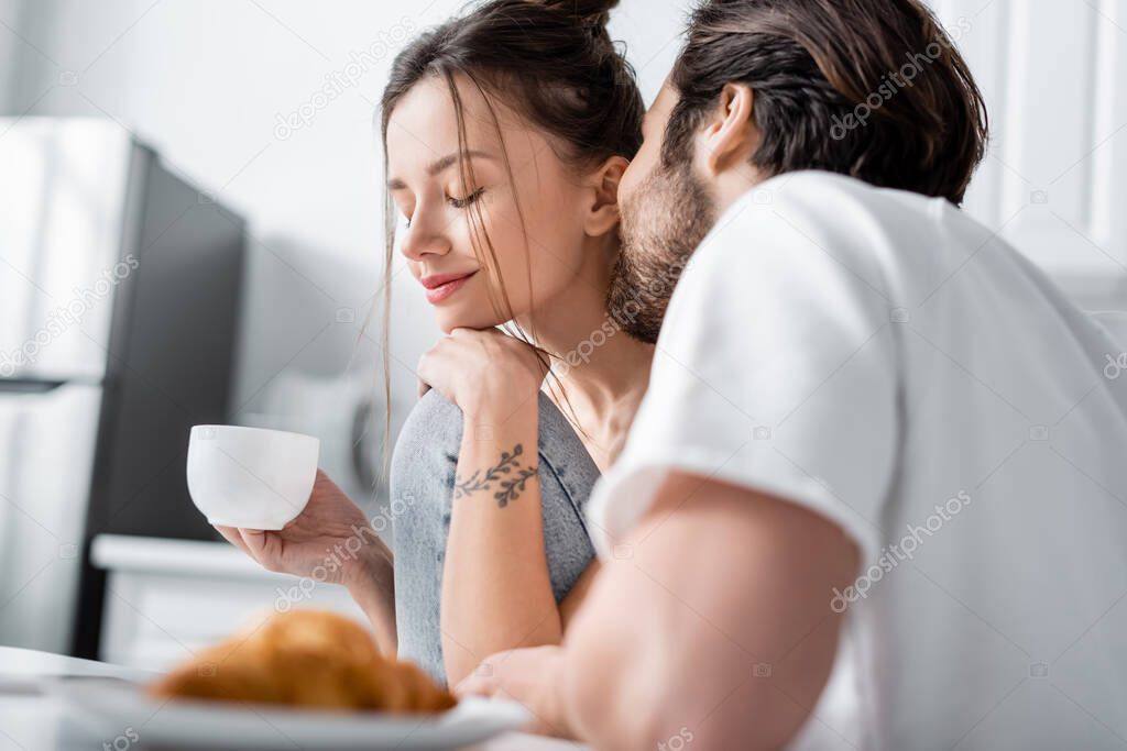 bearded man kissing cheek of tattooed girlfriend with cup of coffee