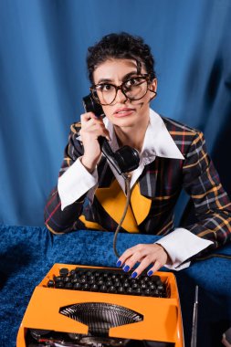 Tedirgin olmuş eski moda haber spikeri kameraya bakarken telefonda mavi arka planda daktilo başında konuşuyor.