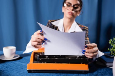 Gözlüklü bulanık bir kadın mavi arka planda daktilonun yanında boş bir kağıt tutuyor.