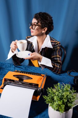 stylish journalist sitting with coffee cup near vintage typewriter on blue background clipart