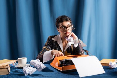 thoughtful newswoman looking away near crumpled paper and vintage typewriter on blue background clipart