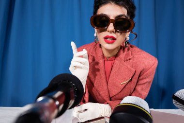 Klasik güneş gözlüklü ve beyaz eldivenli bir kadın mavi arka plandaki görüşme sırasında parmağıyla işaret ediyor. 