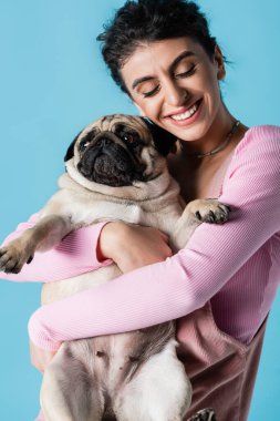 Kapalı gözleri olan neşeli bir kadın. Mavide izole edilmiş komik bir köpeğe sarılıyor.