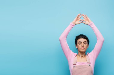 Gözleri kapalı genç bir kadın ellerini kaldırarak meditasyon yapıyor ve jana mudra jesti mavi üzerine izole edilmiş.