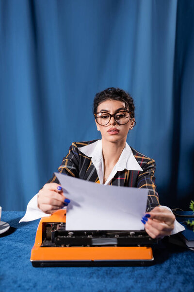 журналист в винтажной одежде держит пустую бумагу возле пишущей машинки на синем фоне