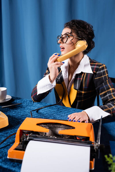 surprised journalist talking on telephone near typewriter on blue background with drapery