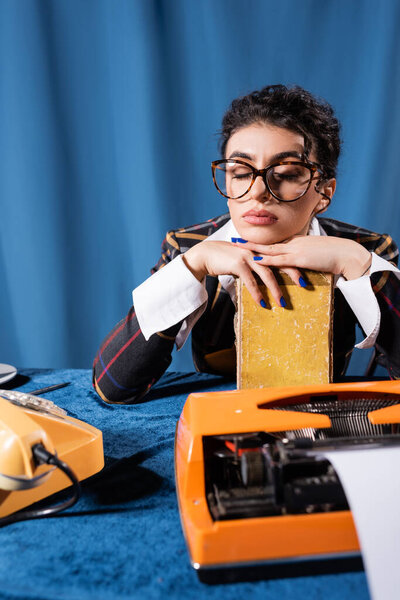 усталая женщина сидит с книгой рядом с винтажной пишущей машинкой и телефоном на синем фоне