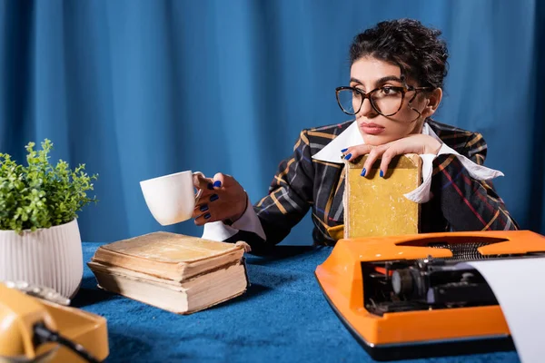 沉思的女人坐在蓝色背景的老式打字机旁 手里拿着咖啡杯和书本 — 图库照片