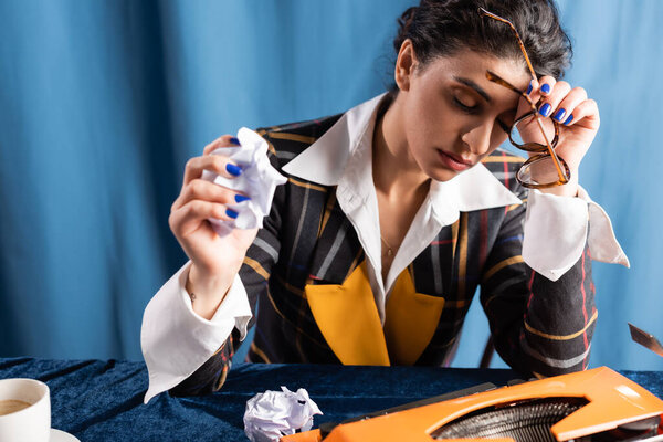 tired journalist holding crumpled paper and eyeglasses while sitting near typewriter on blue background