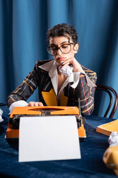 sad newswoman sitting near vintage typewriter with empty paper sheet on blue background