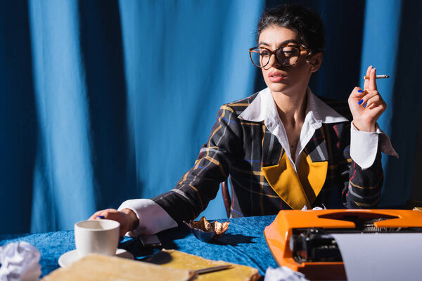 vintage style newswoman with cigarette near coffee cup and typewriter on blue background