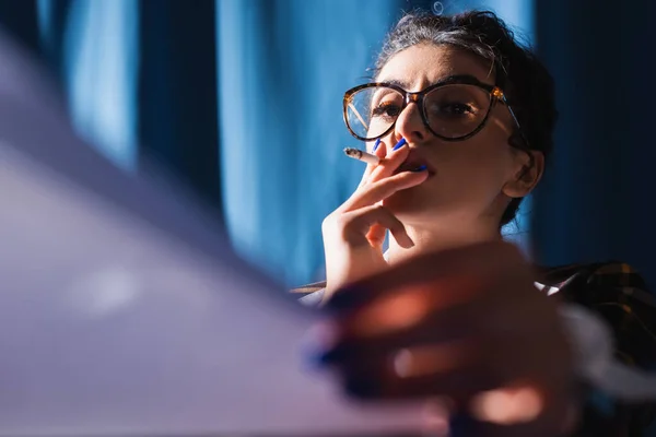 woman in vintage eyeglasses looking at blurred paper while smoking on blue background