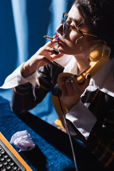 青い背景色の紙の近くでタバコを吸いながら携帯電話を持っているレトロなスタイルの女性 — ストック写真