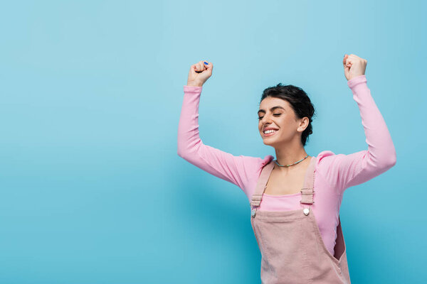 cheerful and stylish woman with closed eyes showing triumph gesture on blue background