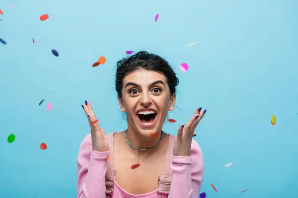 overjoyed woman screaming and showing wow gesture under falling confetti isolated on blue