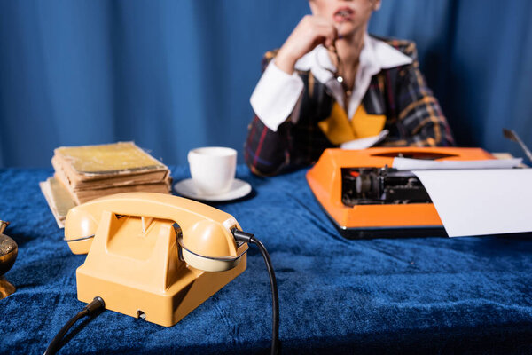 partial view of blurred newswoman near vintage telephone, typewriter and books on blue velour tablecloth