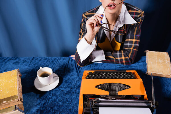 partial view of vintage style woman holding eyeglasses near typewriter and coffee cup on blue background