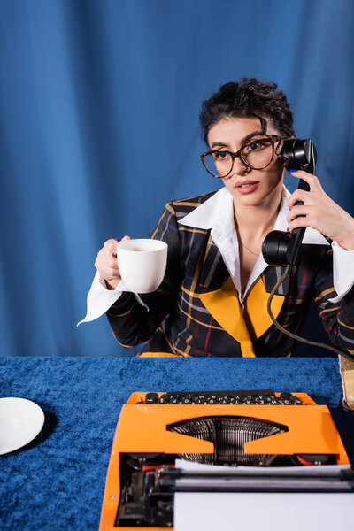 vintage style newswoman with handset and coffee cup near typewriter on blue background