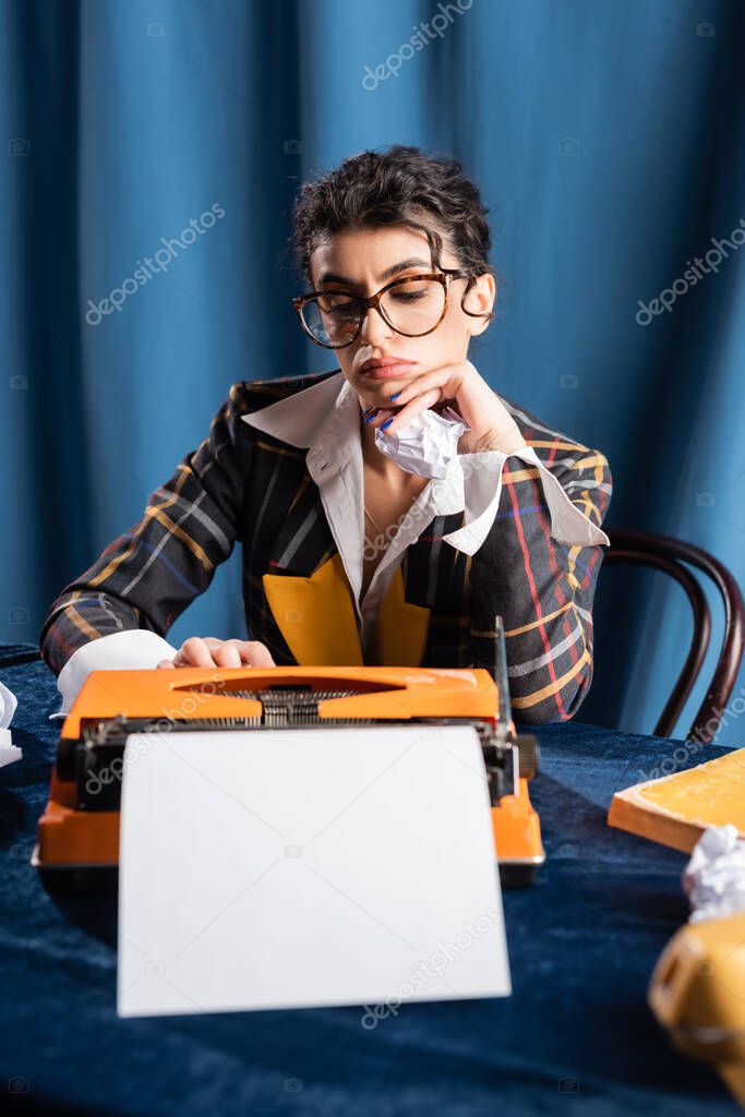 sad newswoman sitting near vintage typewriter with empty paper sheet on blue background