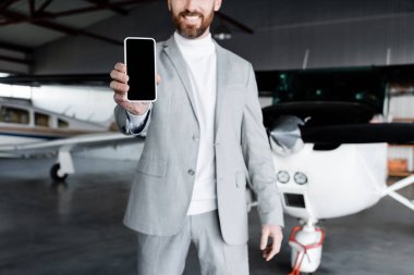 Modern helikopterin yanında boş ekranlı akıllı telefon gösteren resmi giyimli neşeli iş adamının kırpılmış görüntüsü