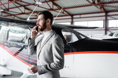 Resmi kıyafetli neşeli bir adam modern helikopterin yanında akıllı telefondan konuşuyor.