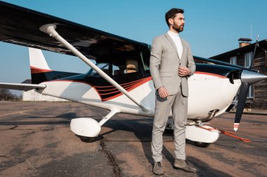 Takım elbiseli sakallı bir iş adamı modern helikopterin yanında dikiliyor ve spor ceketi ayarlıyor.