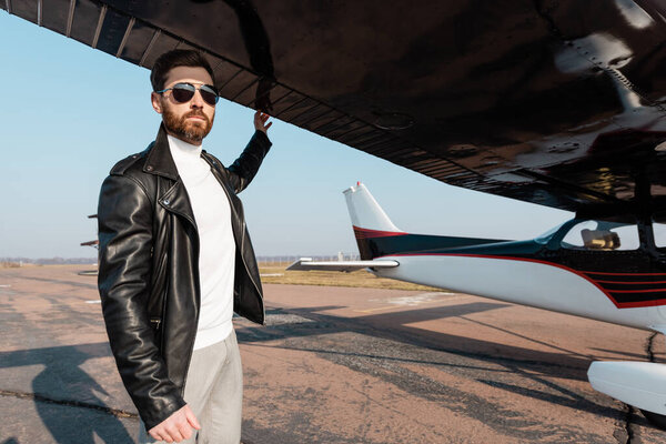 бородатый пилот в солнцезащитных очках и кожаной куртке стоит под крылом самолета