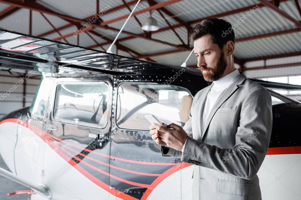 bearded man in formal wear messaging on smartphone near modern helicopter