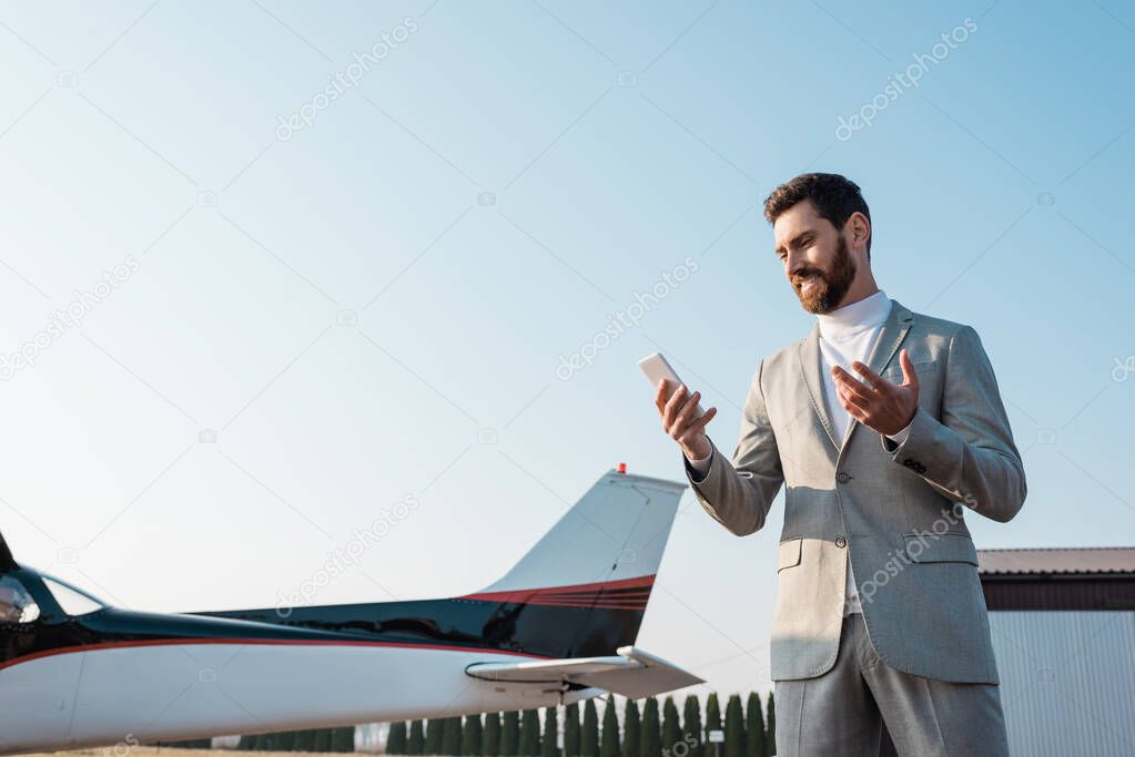 bearded businessman smiling while looking at smartphone near helicopter outdoors 