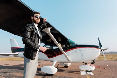 Güneş gözlüklü ve deri ceketli sakallı pilot uçak kanadının yanında duruyor. 