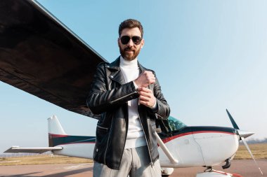 Güneş gözlüklü ve deri ceketli neşeli pilot uçağın yanında duruyor. 