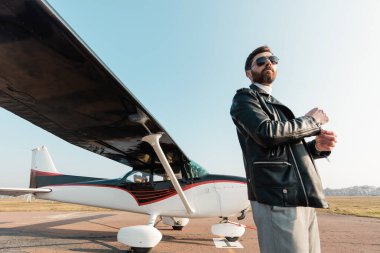 Güneş gözlüklü ve deri ceketli pilotun alçak açılı görüntüsü uçağın yanında duruyor. 