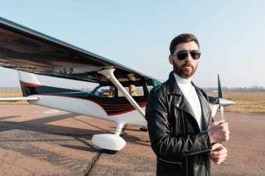 Deri ceketli ve güneş gözlüklü sakallı pilot uçağın yanında duruyor. 
