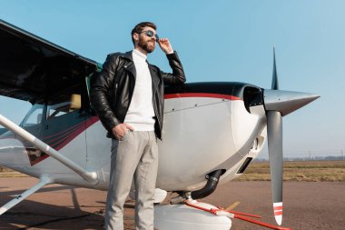 Deri ceketli, güneş gözlüklü ve cebinde uçağın yanında duran mutlu pilotun düşük açılı görüntüsü. 