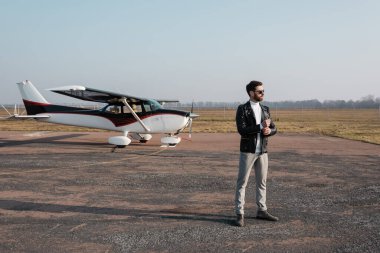Deri ceketli ve güneş gözlüklü şık pilot helikopterin yanında duruyor. 