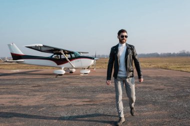 Şık deri ceketli ve güneş gözlüklü mutlu pilot helikopterin yanında yürüyor.