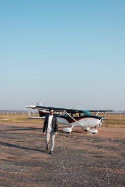 Deri ceketli, güneş gözlüklü ve helikopterin yanında yürüyen memnun bir pilot.