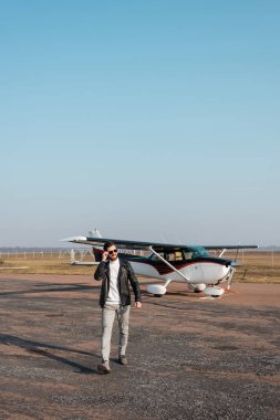 Lüks deri ceketli, helikopterin yanında yürüyen ve güneş gözlüğünü ayarlayan memnun bir pilot.