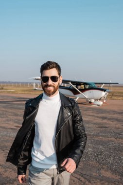 Helikopterin yanında yürüyen şık deri ceketli ve güneş gözlüklü pozitif pilot.