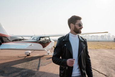 Şık deri ceketli ve helikopterin yanında güneş gözlüklü bir pilot.