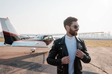 Güneş gözlüklü mutlu pilot helikopterin yanında şık bir deri ceket ayarlıyor.