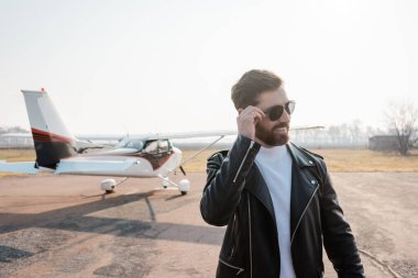 Havalı deri ceketli, helikopterin yanında güneş gözlüğü takan memnun pilot.