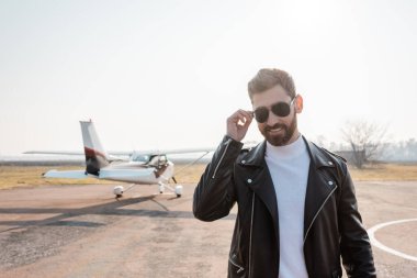 Siyah deri ceketli mutlu pilot helikopterin yanında güneş gözlüğünü ayarlıyor.