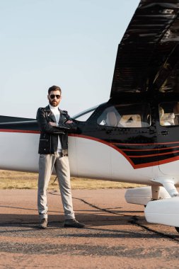 Şık güneş gözlüklü ve deri ceketli sakallı bir pilot uçak kanadının yanında çapraz kollarıyla ayakta duruyor. 