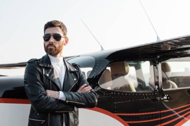 Güneş gözlüklü ve deri ceketli sakallı pilot helikopterin yanında duruyor. 
