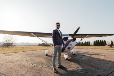 Modern bir uçağın yanında duran şık güneş gözlüklü ve deri ceketli sakallı bir pilot. 