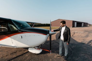 Modern uçaklara dokunan güneş gözlüklü ve deri ceketli sakallı bir pilot. 